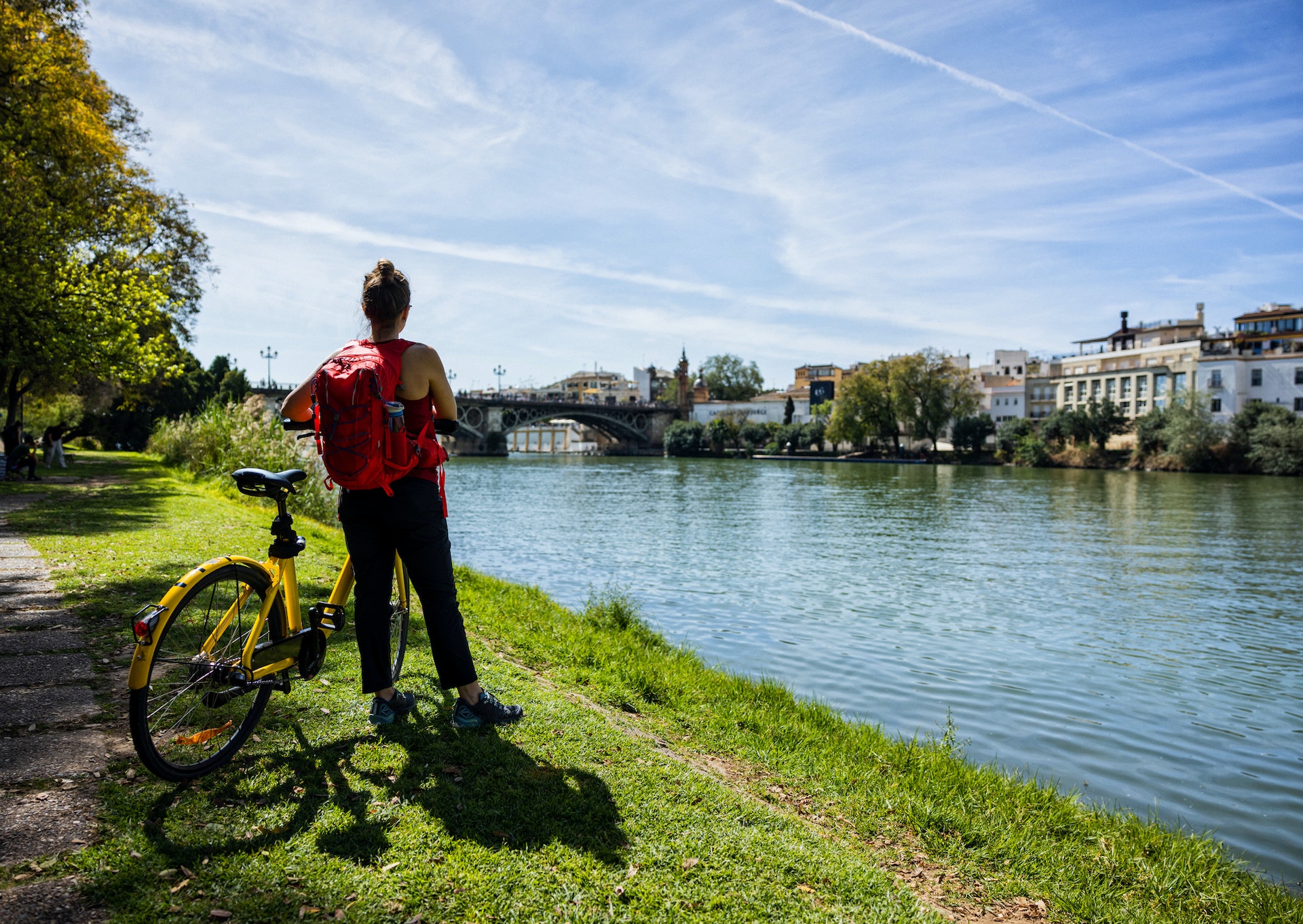 A tourist stands by a river beside her bike looking across at the city skyline on a sunny day